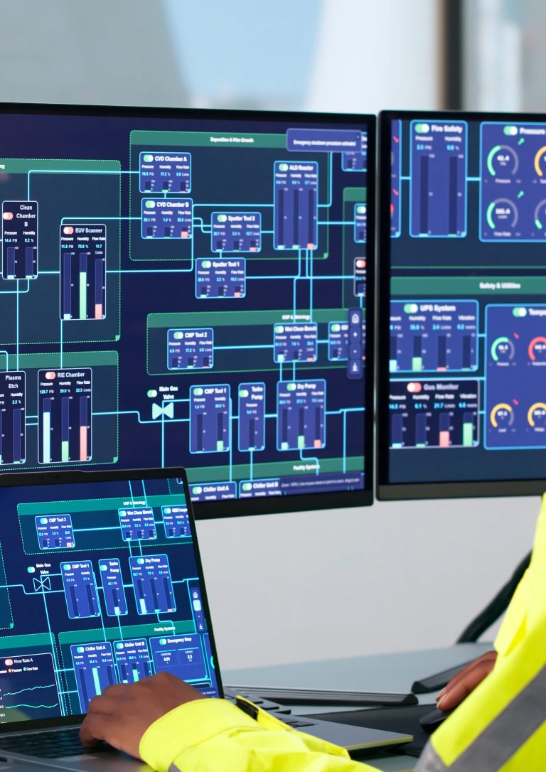 An operator's hands on a laptop and mouse at a control-room desk, with two monitors showing grid system diagrams and gauges.
